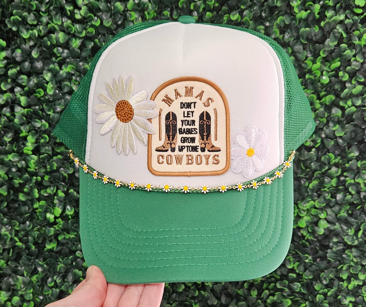 A green and white trucker hat with floral patches and a text patch that reads 'WANIS COWBOYS' on the front. The hat features a snap back closure and a chain accessory around the base.