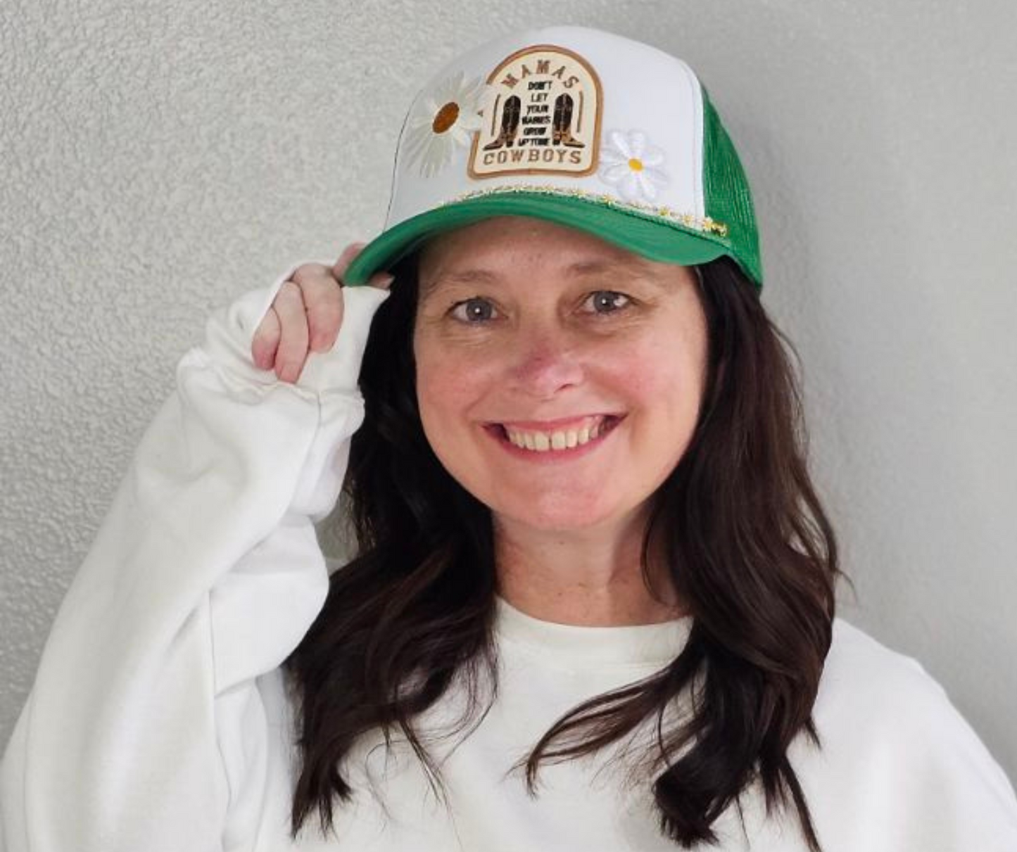 A woman wearing a green and white trucker hat with a 'Mama Don't Let Your Babies Grow Up To Be Cowboys' patch and 2 daisy patches on the front. Daisy chain is also attached to the front of hat.