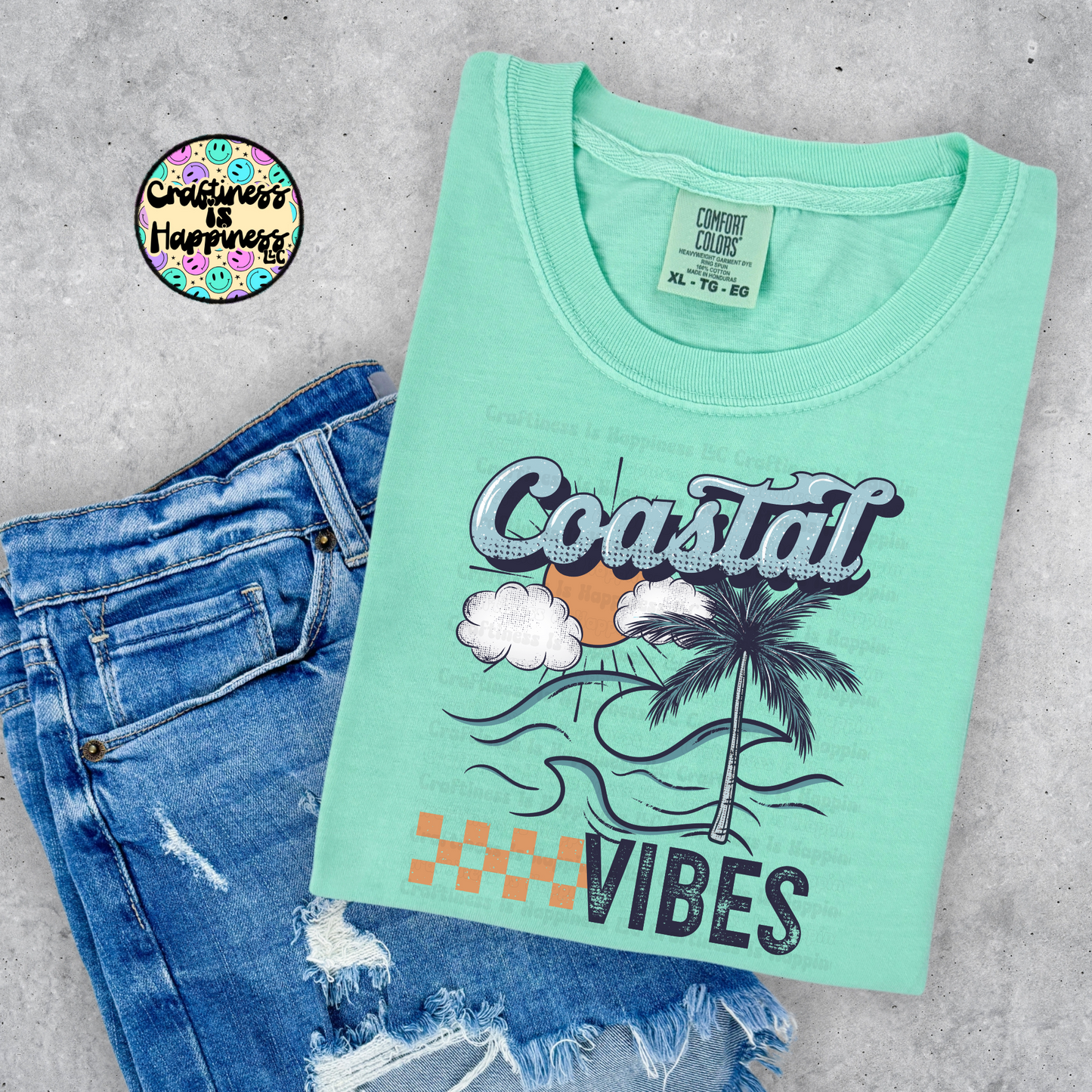 Mint green t-shirt with 'Coastal Vibes' design and blue jeans on a gray surface, featuring a brand logo in the corner.