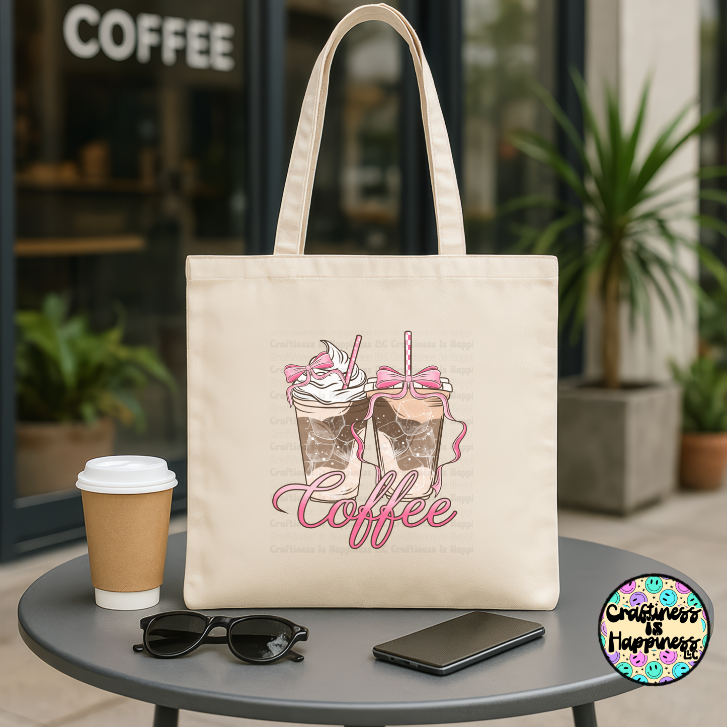 Beige tote bag with coffee design, cup, sunglasses, and phone on a table outside a coffee shop.