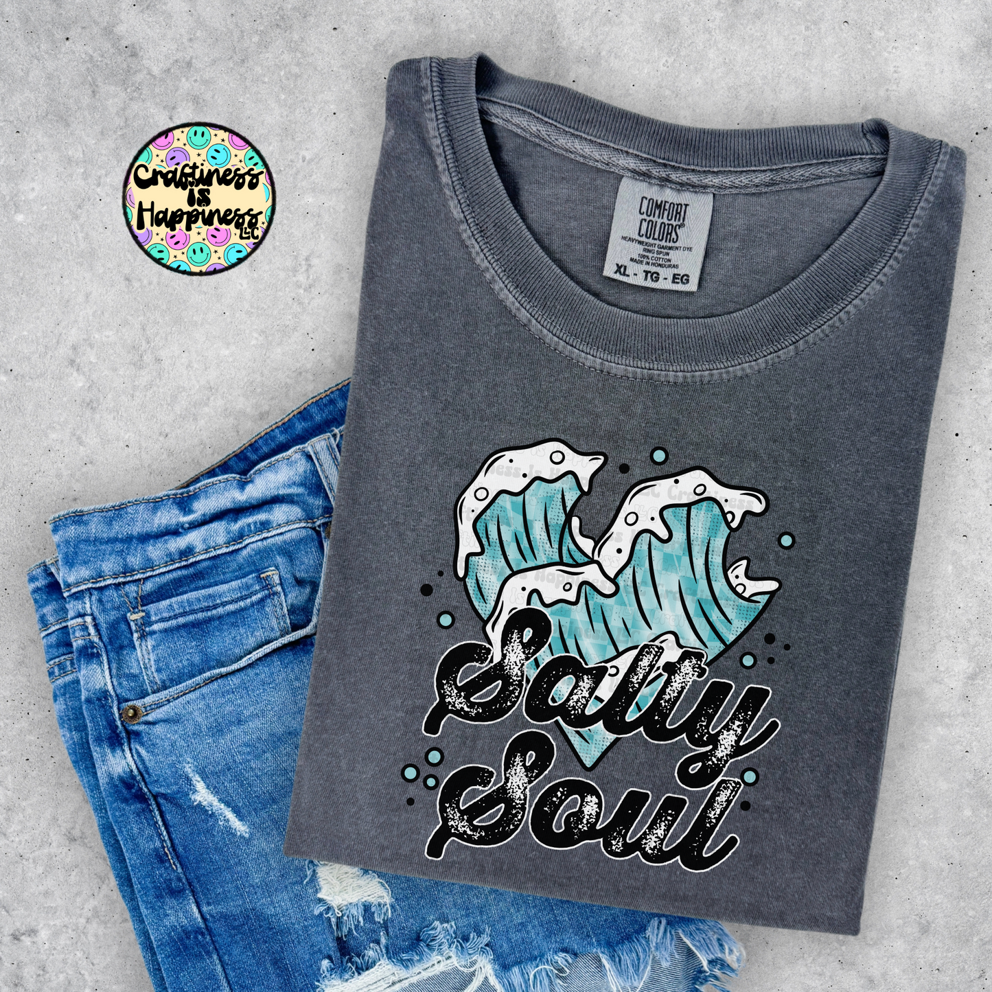 Gray t-shirt with wave design and 'Salty Soul' text, paired with blue jeans on a gray surface.