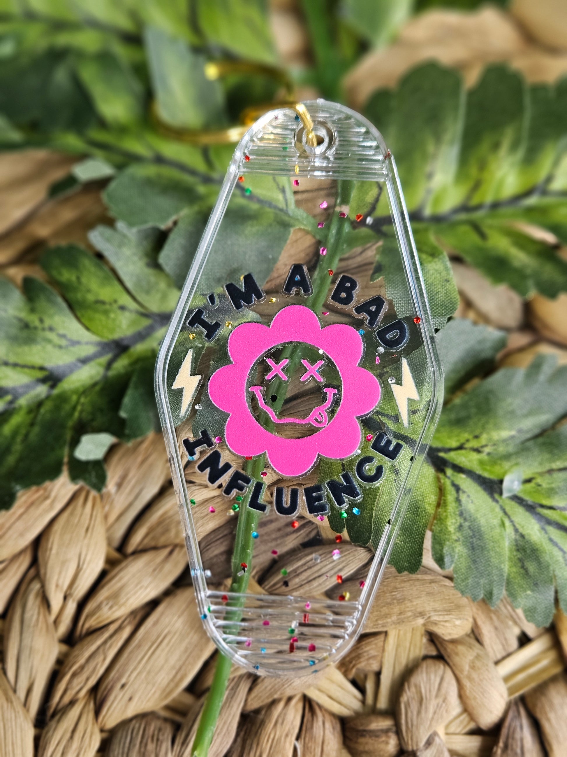 Clear acrylic motel/ retro style keychain with confetti specks . Decorated with a UV DTF sticker with black text "I'm A Bad Influence" with a pink retro daisy smiley face, and yellow lightning bolt on each side of daisy.