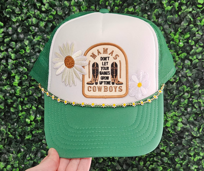 A green and white trucker hat with floral patches and a text patch that reads 'WANIS COWBOYS' on the front. The hat features a snap back closure and a chain accessory around the base.