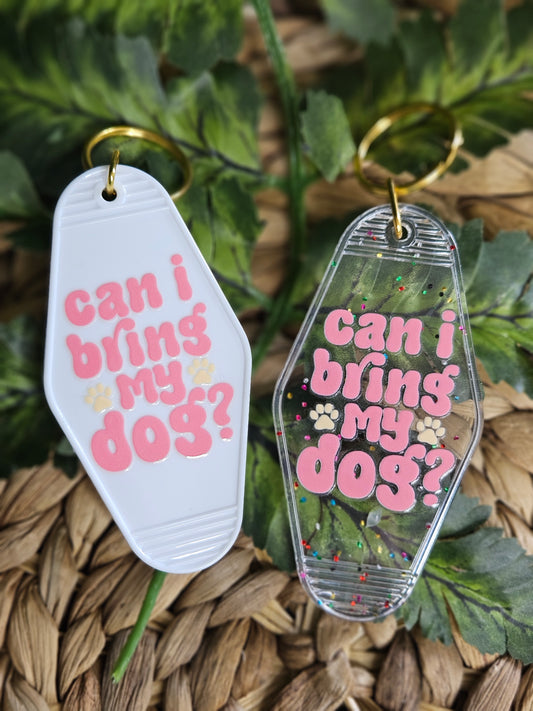 Available in a plain white or clear acrylic motel/ retro style keychain with confetti speckles. Decorated with a pink retro font UV DTF Decal" Can i bring my dog". One cream colored paw print is on each side of the word "my". Both Keychain color options have gold colored hardware.