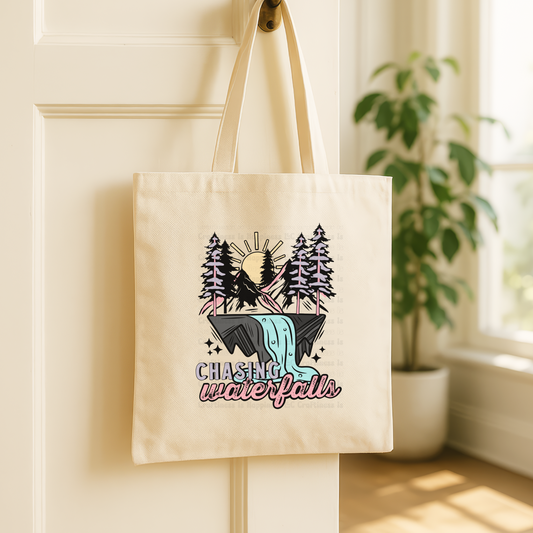 Beige tote bag with nature-themed design and text 'Chasing Waterfalls' hanging on a white wall.