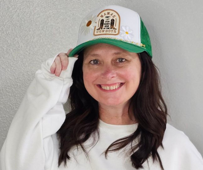 A woman wearing a green and white trucker hat with a 'Mama Don't Let Your Babies Grow Up To Be Cowboys' patch and 2 daisy patches on the front. Daisy chain is also attached to the front of hat.