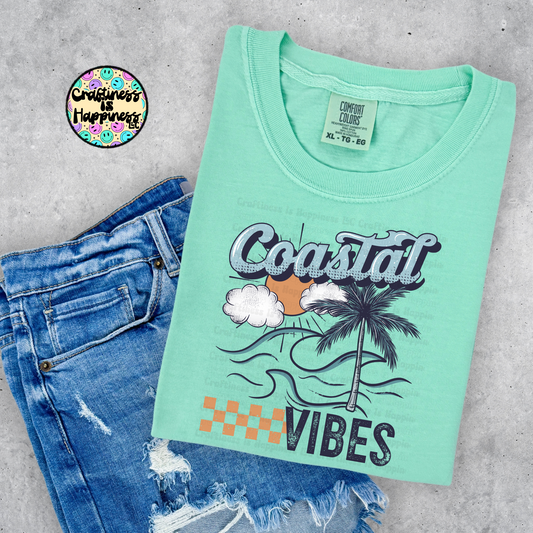 Mint green t-shirt with 'Coastal Vibes' design and blue jeans on a gray surface, featuring a brand logo in the corner.