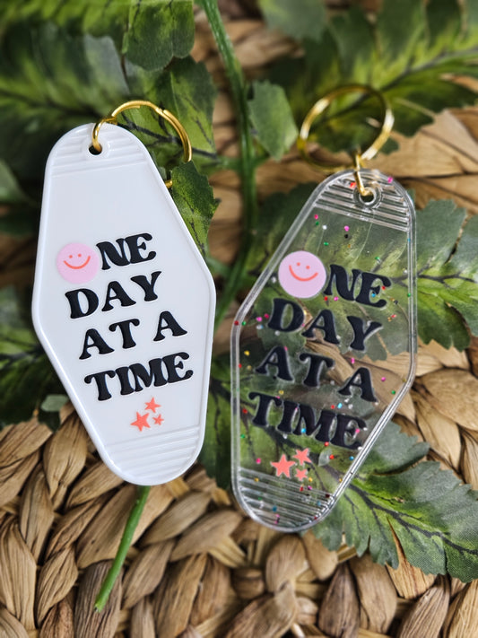 Available in a plain white or clear acrylic motel/ retro style keychain with confetti speckles. Decorated with a black font UV DTF Decal" One day at a time". A pink smiley face is in the place of the letter "o" in the word one, with 3 coral/ orange stars in various sizes located under the word time. Keychains also have gold colored hardware.