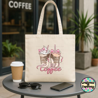 Beige tote bag with coffee design, cup, sunglasses, and phone on a table outside a coffee shop.