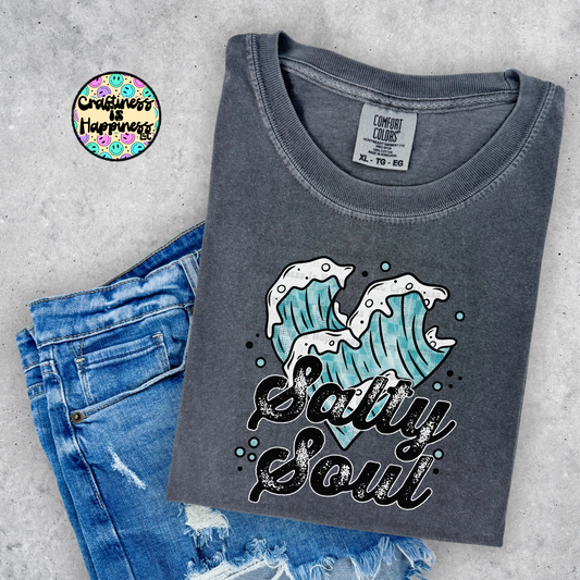 Gray t-shirt with wave design and 'Salty Soul' text, paired with blue jeans on a gray surface.