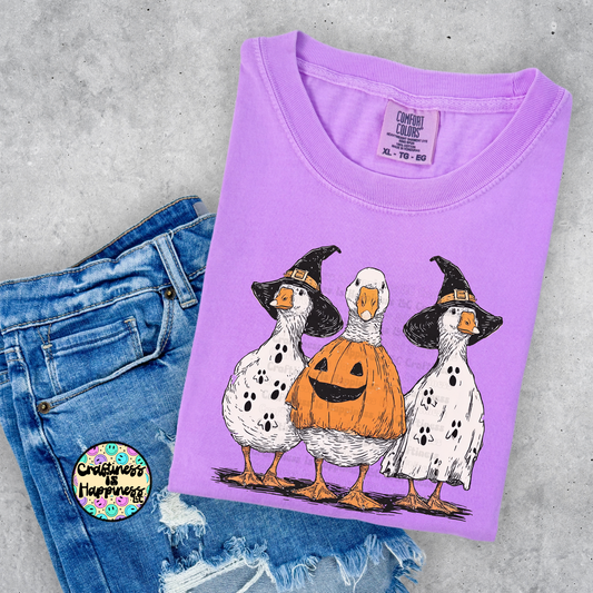 Purple t-shirt with Halloween-themed duck design on a concrete surface with denim shorts.